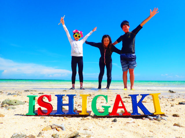 石垣島1泊2日旅行の記念写真。カラフルな「ISHIGAKI」のロゴと、晴天のビーチで楽しむ家族。